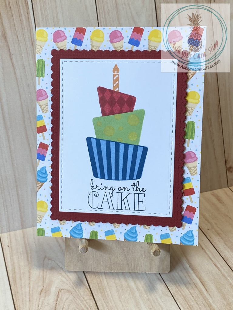 Brightly coloured birthday themed patterned paper background of ice cream. A solid coloured scalloped panel in red is popped up on the centre of the card. On top is a smaller white card panel with faux stitched detail stamped with a multi-tiered crooked birthday cake, also brightly coloured. The sentiment reads: bring on the cake. Internal sentiment reads: don't count your candles count your blessings. Card size is A2 with coordinating white envelope.