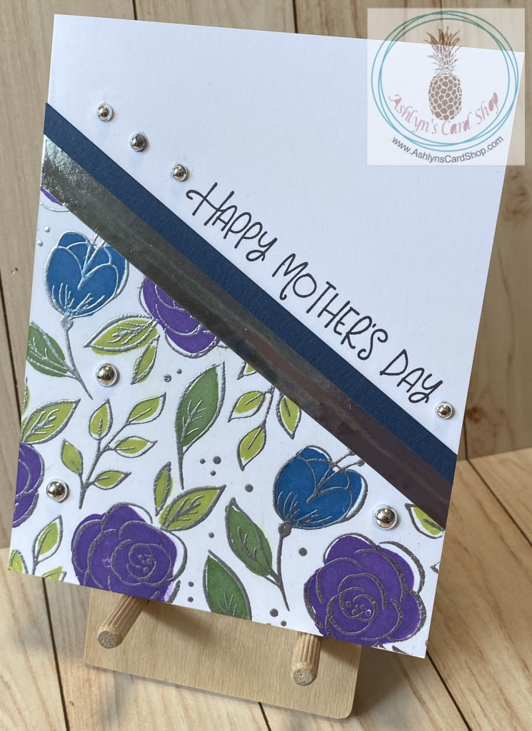 A simple one layer card with a stamped and embossed floral design on the bottom left. Coloured cardstock and a metallic stripe complement the floral design. Metallic bubbles are added as an accent. External sentiment reads "Happy Mother's Day" and the internal sentiment reads "I am a strong woman because I was raised by a strong woman." Blue & silver version. Card size is A2 (4.5 x 5.5"). Coordinating envelope included.