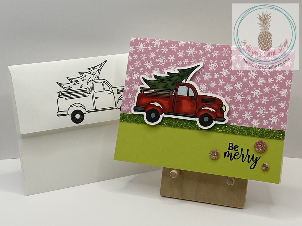 Getting The Christmas Tree Card - A hand coloured image of a truck bringing home a fresh Christmas tree. Available  in two snowy background options: pink and blue. External sentiment reads “Be merry” and the internal sentiment reads “Happy Holidays”.  A2 size card: 4.25 x 5.5”. Coordinating envelope (shown) included.