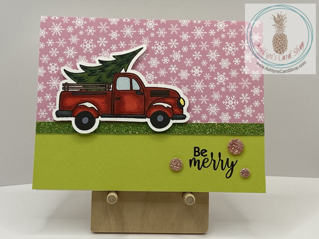 Getting The Christmas Tree Card - A hand coloured image of a truck bringing home a fresh Christmas tree. Available  in two snowy background options: pink (shown) and blue. External sentiment reads “Be merry” and the internal sentiment reads “Happy Holidays”.  A2 size card: 4.25 x 5.5”. Coordinating envelope included.