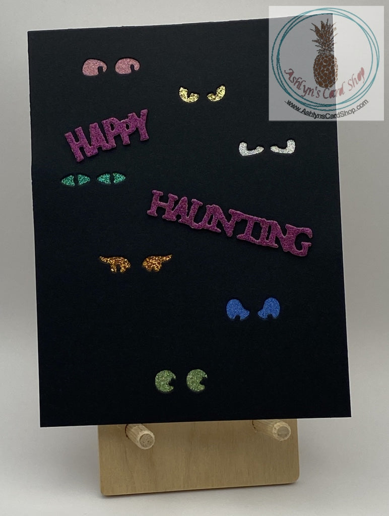 A black cardstock background with spooky glittery eyes in multiple colours peering out! External sentiment reads: happy haunting; inside is blank for your personal message. A2 size card (4.25 x 5.5"). Coordinating envelope included.
