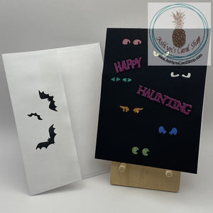 A black cardstock background with spooky glittery eyes in multiple colours peering out! External sentiment reads: happy haunting; inside is blank for your personal message. A2 size card (4.25 x 5.5"). Coordinating envelope (shown) included.