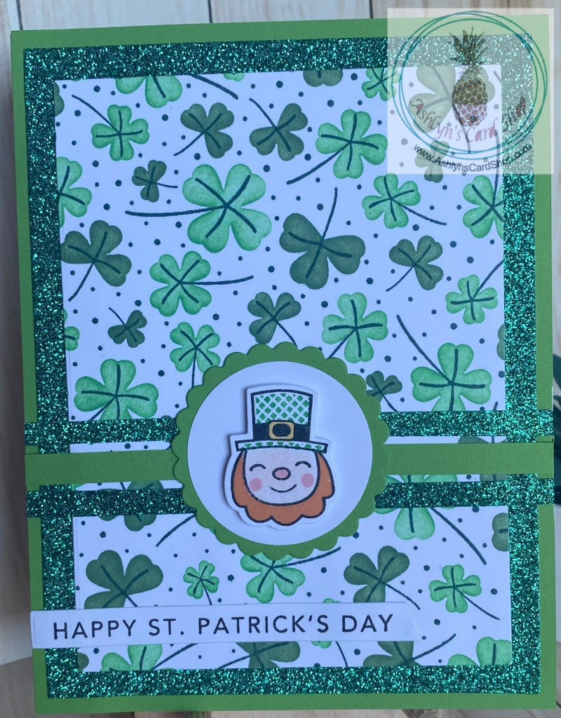 A handmade greeting card to celebrate St. Patrick’s Day! The three and four leaf clover background is ink blended using shades of green.  A sparkly green border is added for some interest and that panel is attached to a green bright green panel.  The card front is decorated with a scalloped circle and a cheerful leprechaun face popped up on the centre.  Green and green sparkle stripes provide an accent across the card behind the scalloped circle. A simple sentiment (Happy St. Patrick’s Day) on a white senti