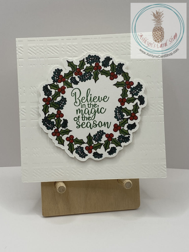 A green wreath with red berries popped up on a white embossed background. Traditional colours used on this Christmas card. External sentiment reads “Believe in the magic of the season” and the internal sentiment reads “have a holly jolly Christmas.” 4” square card.  Coordinating envelope included.