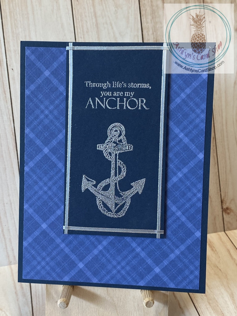Masculine birthday card with blue plaid patterned paper on a navy blue card panel.  A navy blue cardstock panel has an embossed anchor and the sentiment"through life's storms, you are my anchor," with silver metallic lines framing the panel. The panel is popped up in the centre of the top of the card. Internal sentiment reads: happy birthday. Card size is 4.25 x 5.5" (A2). Coordinating white envelope included.