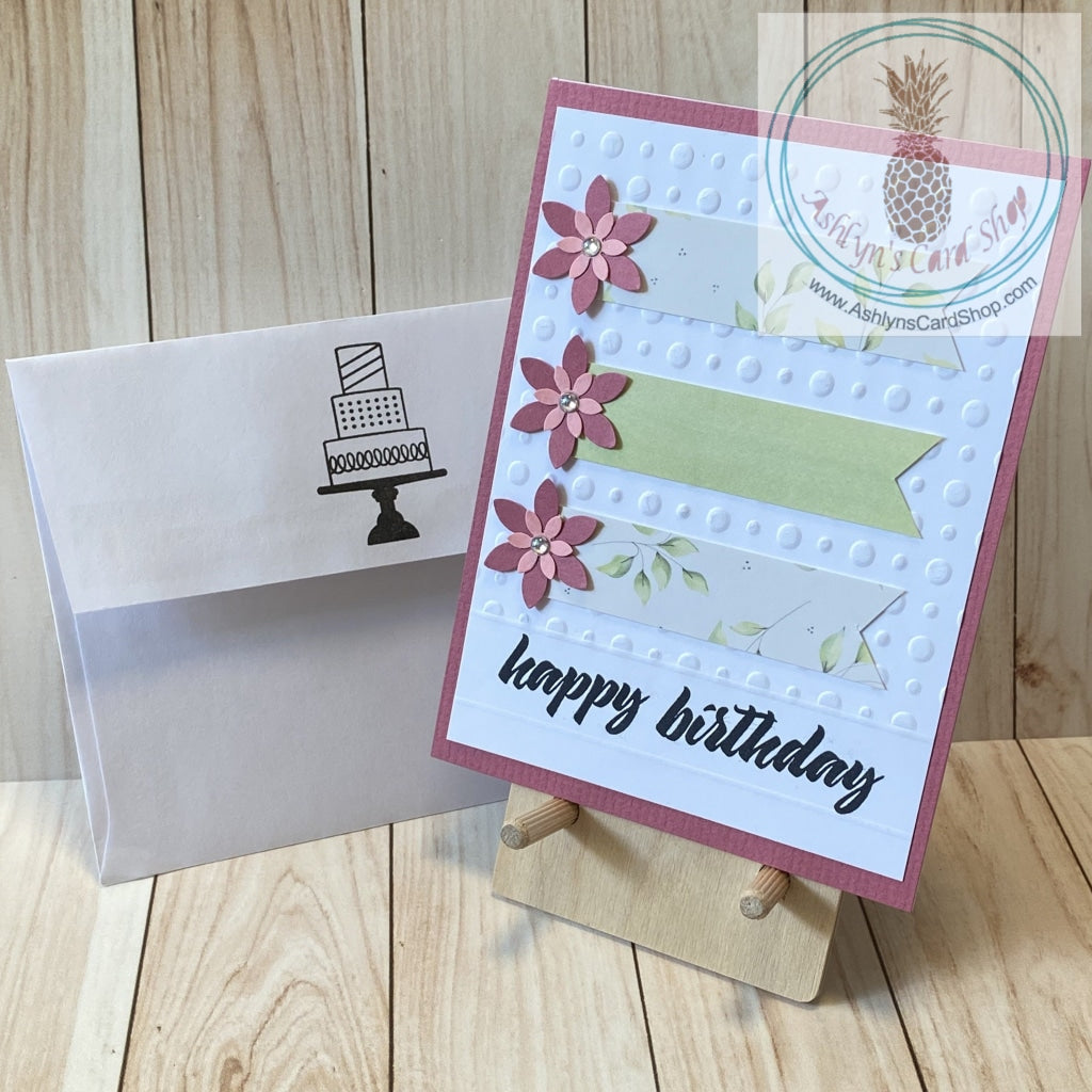 High quality handmade greeting card featuring layered pink flowers and banners of coordinating patterned paper on white card panel embossed with dots and stamped with "happy birthday". The white panel is attached to a pink cardstock background on a white card base.  Internal sentiment reads "make a wish". Green version. A2 card size: 4.25 x 5.5".  Coordinating envelope (shown) included.