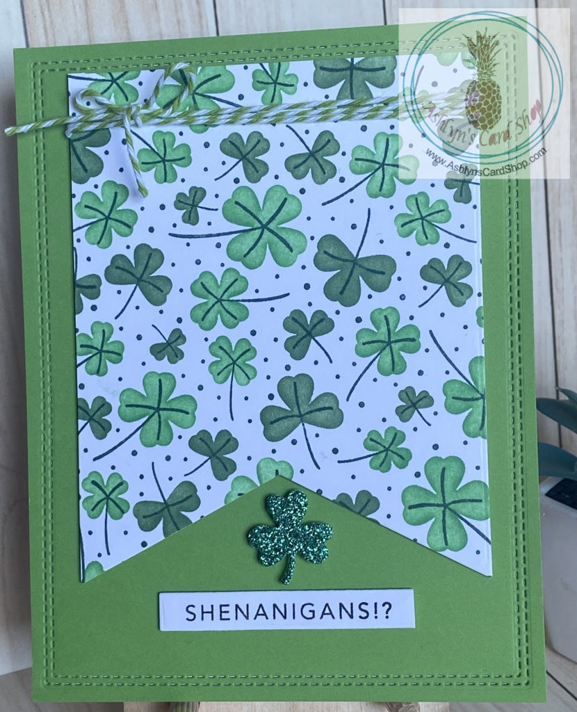A handmade card in shades of green to celebrate St. Patrick’s Day! Know someone who engages in shenanigans? Faux stitched detail around the outer edge of the card panel.  A white banner with shamrocks ink blended in shades of green is popped up on the front with a twine bow accent at the top.  A green sparkly shamrock and the “shenanigans” sentiment complete the card design.  Internal sentiment reads: happy St. Patrick’s Day.  Card size: 4.25 x 5.5” (A2)  Coordinating envelope included.