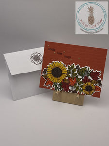 Hand coloured fall floral collage on an embossed orange background. External sentiment reads: smile love hugs; inside of card is blank for your personal message. A2 size card 4.25 x 5.5". Coordinating envelope (shown) included.
